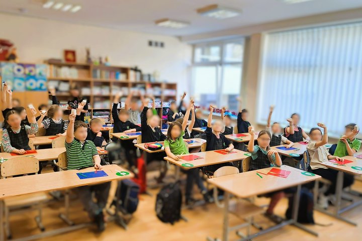 Pradinukai klasėje sėdi prie suolų ir iškėlę rankas laiko balsavimo korteles ar ženklus, klasėje tvyro aktyvi ir įsitraukusi atmosfera, fone matyti lentynos su knygomis ir mokymo priemonėmis.