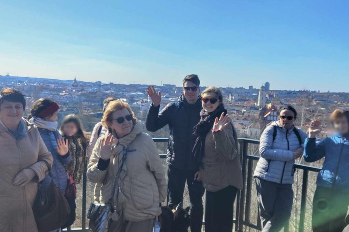 Grupė žmonių sveikinasi mojuodami nuo apžvalgos aikštelės su vaizdu į Vilniaus senamiestį. Fone matyti Šv. Jonų bažnyčios bokštas ir kiti senamiesčio pastatai. Nuotraukoje matyti giedras, saulėtas oras.