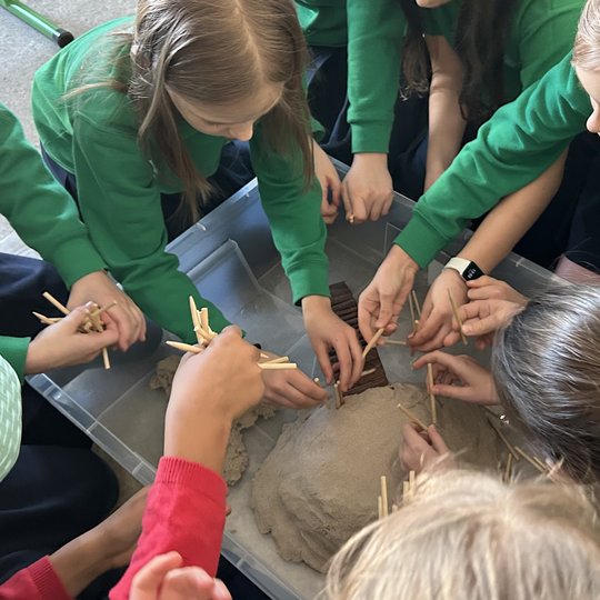Grupė vaikų su žaliomis uniformomis susibūrę aplink plastikinę dėžę su smėliu, kurioje formuoja piliakalnio ar tvirtovės maketą, naudodamiesi mediniais pagaliukais ir kitomis priemonėmis, aktyviai dalyvauja kūrybinėje edukacinėje veikloje.