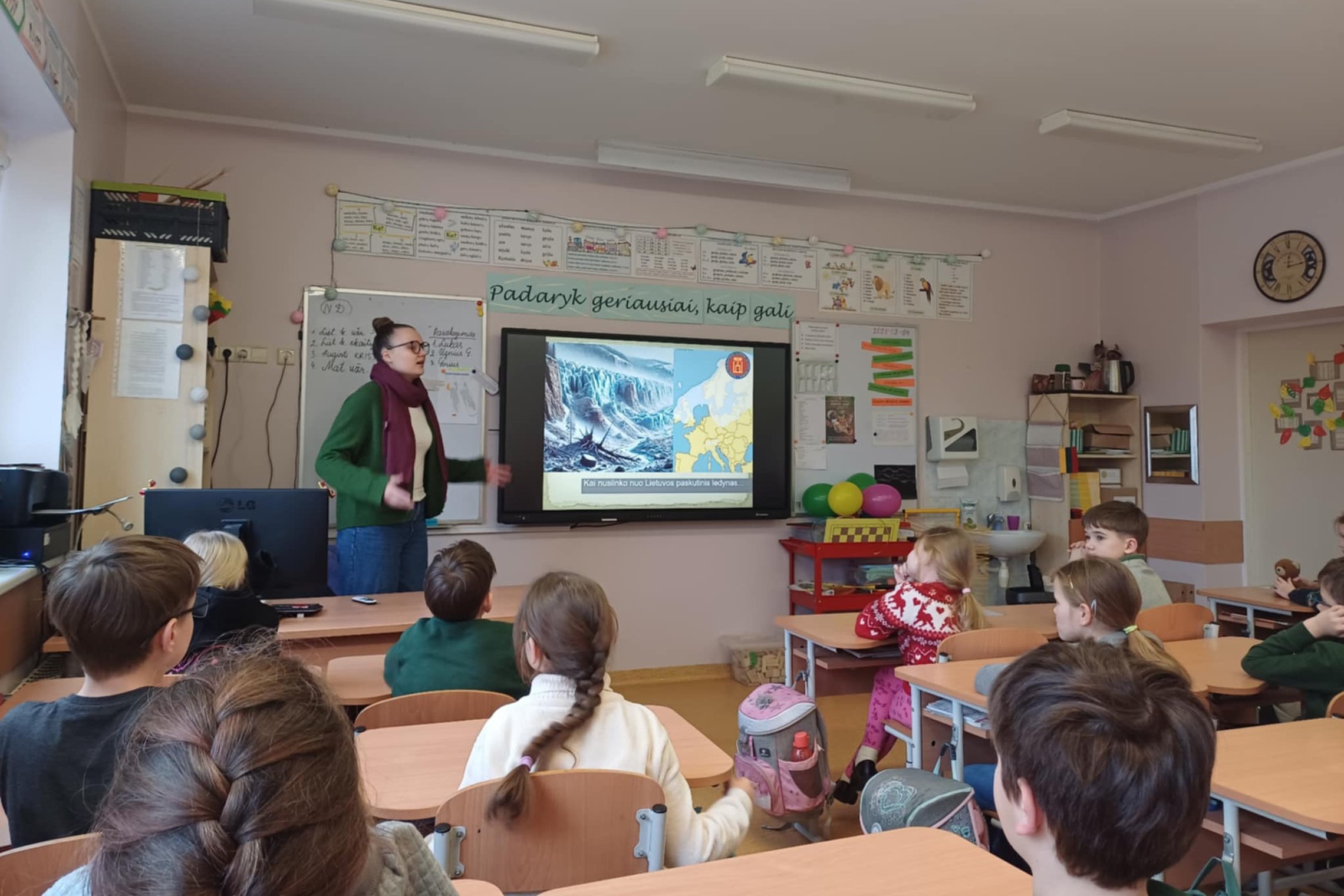 Šioje nuotraukoje užfiksuota edukacinė pamoka pradinukų klasėje. Priekyje stovi edukatorė, pasakojanti vaikams apie geologinius ar klimato reiškinius – skaidrėje matyti tema „Kai nuslinko nuo Lietuvos paskutinis ledynas“, o šalia pateiktas žemėlapis bei l
