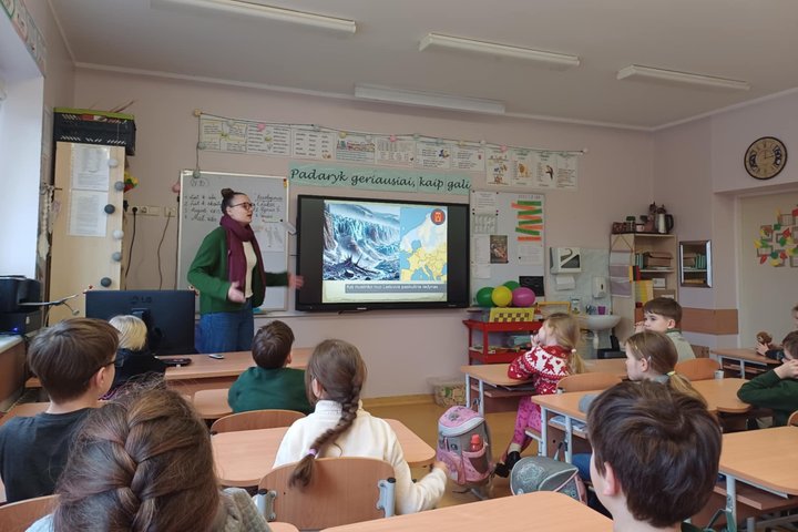 Šioje nuotraukoje užfiksuota edukacinė pamoka pradinukų klasėje. Priekyje stovi edukatorė, pasakojanti vaikams apie geologinius ar klimato reiškinius – skaidrėje matyti tema „Kai nuslinko nuo Lietuvos paskutinis ledynas“, o šalia pateiktas žemėlapis bei l