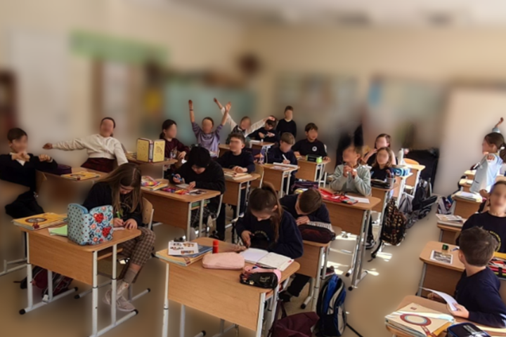 Klasėje prie suolų sėdi įvairia veikla užsiėmę mokiniai – vieni rašo, kiti kelia rankas ar šnekučiuojasi; ant stalų matyti sąsiuviniai, vadovėliai, penalai ir kitos mokymosi priemonės, o fone – šviesi klasės aplinka su lenta ir lentynomis.