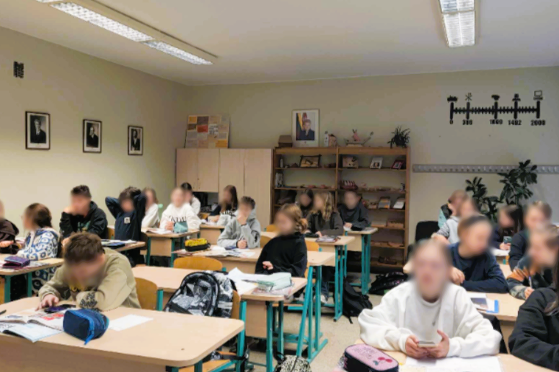 Klasės kambaryje vaikai sėdi prie savo suolų. Kai kurie mokiniai dėmesingai žiūri į savo užrašų knygas ar vadovėlius, kiti naudojasi mobiliaisiais telefonais. Kambarys yra šviesus ir tvarkingas, su knygų spintomis ir mokomaisiais plakatais ant sienų
