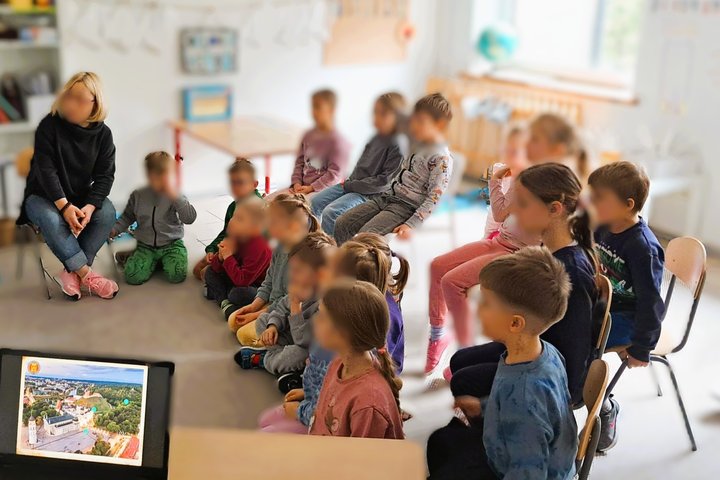 Maži vaikai sėdi puslankiu aplink mokytoją šviesioje klasėje ir žiūri į ekraną, kuriame rodomas Vilniaus pilių vaizdas iš oro su pažymėtais objektais; aplinka jauki, matyti vaikų kūrybos darbai, lentynos, gaublys ir langas su dienos šviesa.