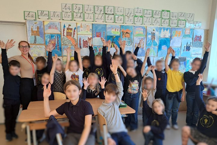 Klasės nuotraukoje matyti gausus būrys mokinių ir edukatorė, visi draugiškai pozuoja iškėlę rankas. Vaikai vilki mokyklines uniformas – dauguma tamsiai mėlynos spalvos, o kai kurie – geltonais marškinėliais ar margais drabužiais su piešiniais. Jie stovi a