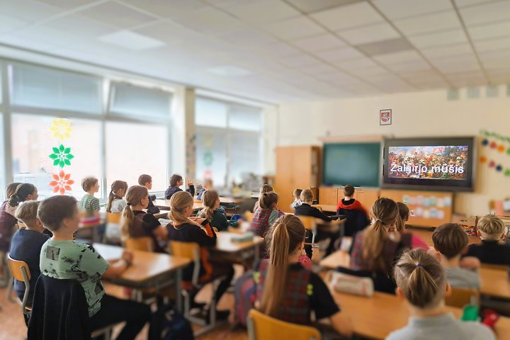 Klasėje sėdintys pradinių klasių mokiniai žiūri vaizdo įrašą apie Žalgirio mūšį. Ant sienos pakabintas televizorius rodo užrašą „Žalgirio mūšis“ su mūšio scena fone. Vaikai sėdi tvarkingai prie stalų, kai kurie vilki languotus uniforminius drabužius. Kamb