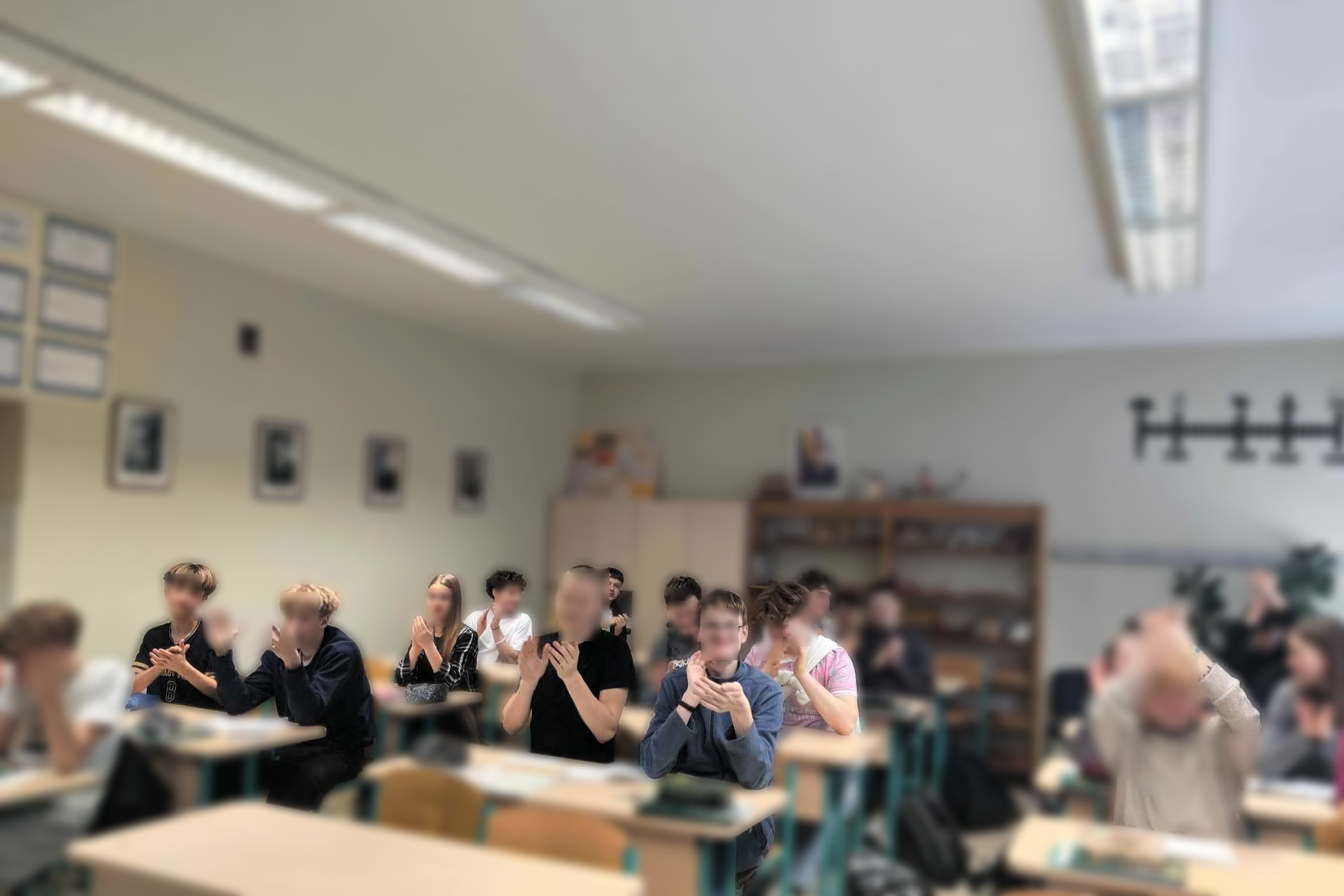 Klasėje prie mokyklinių suolų sėdi grupė paauglių, plojančių rankomis ir šypsantis, lyg reaguojančių į pasirodymą ar pranešimą. Aplink matyti lentynos su mokymo priemonėmis, ant sienų kabo portretai ir kiti mokykliniai dekorai. Nuotaika atrodo džiugi ir į