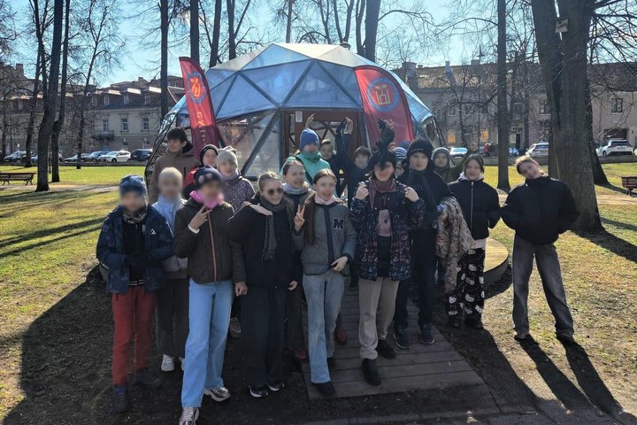 Prie kupolo formos stiklinės struktūros, papuoštos dviem raudonomis vėliavomis su Vilniaus pilių rezervato herbu, stovi grupė vaikų ir paauglių, pozuojančių bendroje nuotraukoje. Aplink parkas su medžiais, šviečia saulė, o fone matyti senamiesčio pastatai