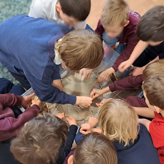 Vaikai iš Filaretų pradinės mokyklos grupiniu darbu stato piliakalnio modelį – smėlio kalne formuoja gynybinę struktūrą naudodami pagaliukus ir figūrėles. Edukacija skatina bendradarbiavimą ir susipažinimą su senovės pilių statybos principais.