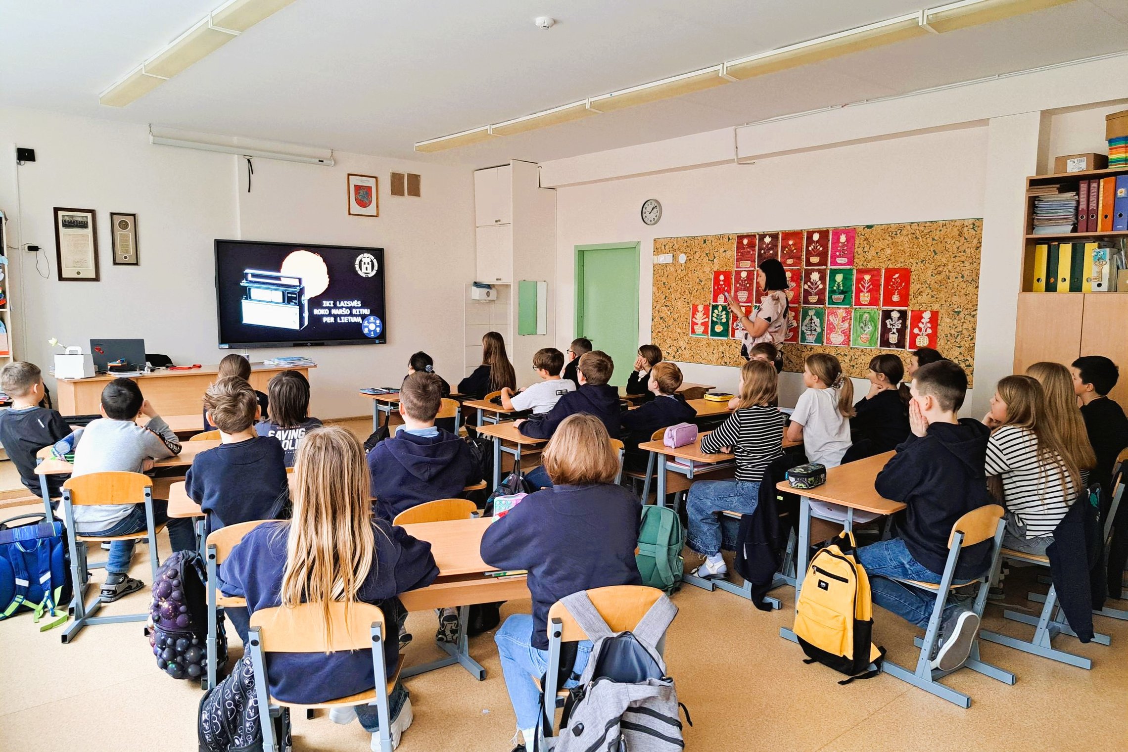 Klasėje mokiniai dalyvauja edukacijoje „Į laisvę roko maršo ritmu per Lietuvą“. Priekyje matomas ekranas su skaidre, kurioje senovinis radijo vaizdas, klasės priekyje stovi edukatorė. Mokiniai sėdi po kelis prie stalų, aplinkoje matyti spalvingi vaikų dar