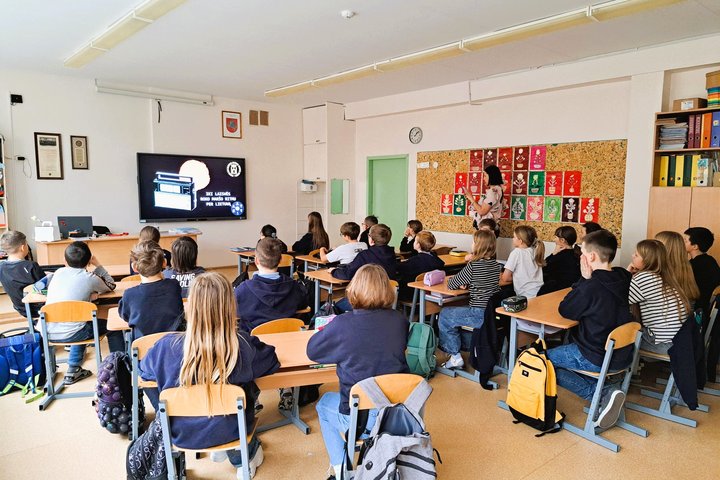 Klasėje mokiniai dalyvauja edukacijoje „Į laisvę roko maršo ritmu per Lietuvą“. Priekyje matomas ekranas su skaidre, kurioje senovinis radijo vaizdas, klasės priekyje stovi edukatorė. Mokiniai sėdi po kelis prie stalų, aplinkoje matyti spalvingi vaikų dar