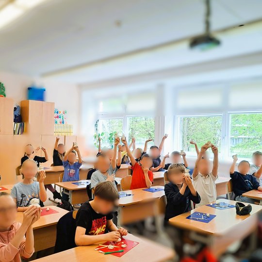 Nuotraukoje matome klasę pilną vaikų. Rankose jie laiko figūrėles. Aplinka šviesi, klasė pilna mokymosi priemonių. Ant stalų yra įrankiai, priemonės