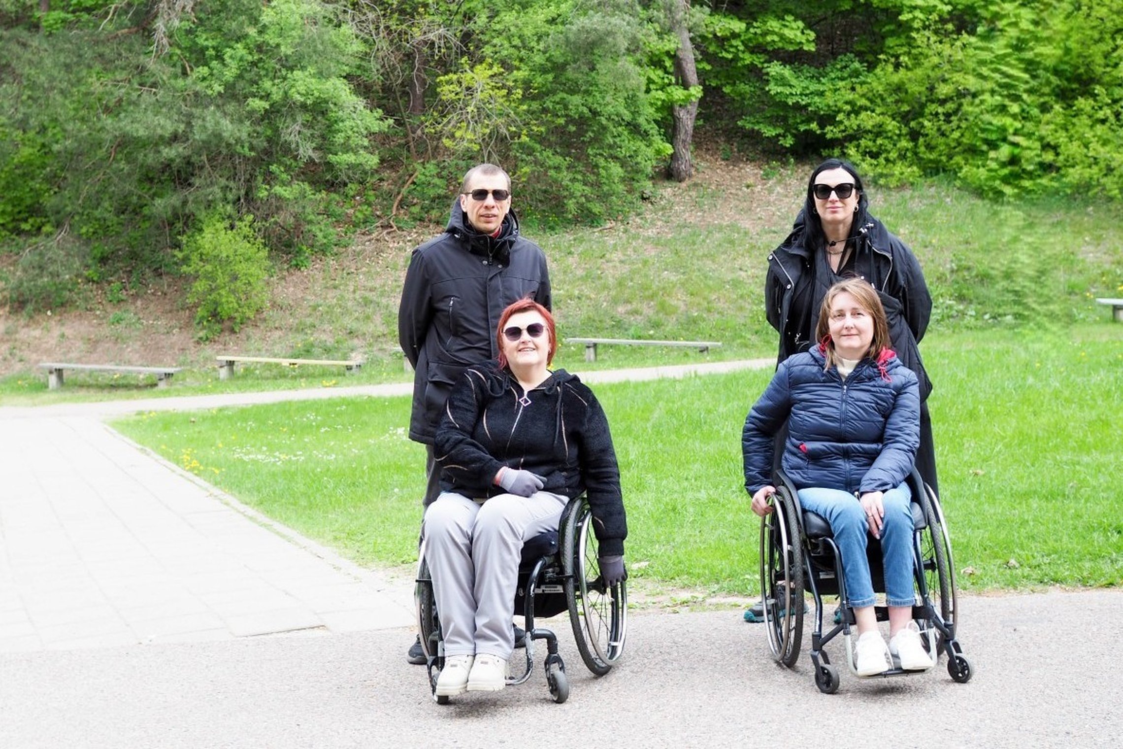 Du žmonės su negalia sėdi neįgaliųjų vežimėliuose, o už jų stovi du asmenys. Visi keturi žmonės šypsosi ir pozuoja Vilniaus pilių teritorijoje, žalioje erdvėje, šalia takelio ir suolelių.