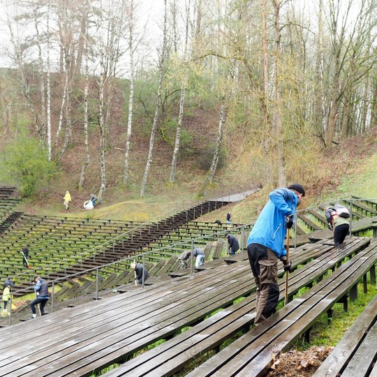 Lauko estradoje ant medinių suoliukų žmonės aktyviai dalyvauja talkoje – valo, grėbia lapus ir renka šiukšles. Priekyje matyti vyras mėlyna striuke, lenkiantis nugarą ir dirbantis su grėbliu. Toliau – daugiau talkininkų, išsibarstę per visą teritoriją.