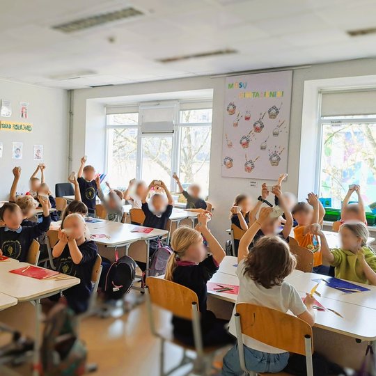 Nuotraukoje matome pradinių klasių mokinius klasėje. Vaikai sėdi prie stalų, pakėlę rankas aukštyn, laikydami kažką rankose – tai modelinas. Klasėje šviesu, pro didelius langus sklinda dienos šviesa. Ant sienų kabinami plakatai, vienas jų – „MŪSŲ GIMTADIE