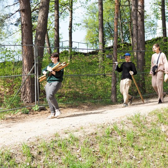 Trys žmonės eina miško taku – vienas neša kuoliukus, kitas turi kastuvą ir kuolą, o trečiasis laiko pirštines. Fone matyti medžiai ir metalinė tvora palei kalvos šlaitą.