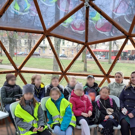 Grupė vaikų ir dvi suaugusios moterys sėdi kupolo formos medinėje konstrukcijoje su skaidriais langais, fone matomas parkas, toliau gatvė ir pravažiuojantis raudonas autobusas.