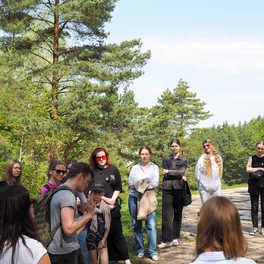 Grupė žmonių stovi puslankiu, klausydamiesi jauno vyro su kuprine, kuris kalba ir gestikuliuoja. Fone matyti spygliuočių miškas ir giedras dangus virš žalios gamtos.
