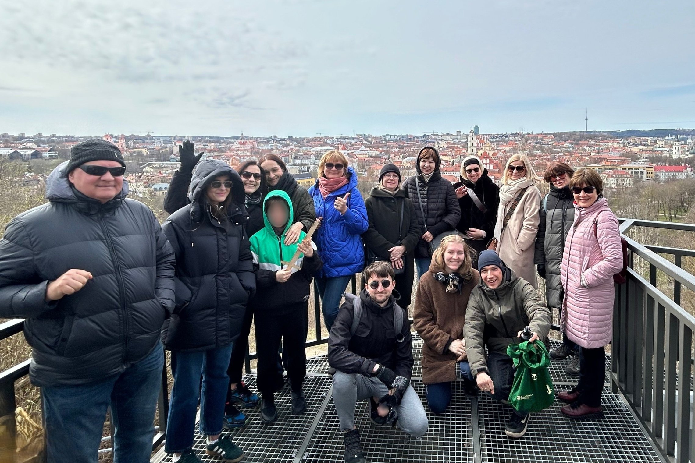 Grupė suaugusiųjų ir vaikų stovi ant Altanos bokšelio terasos, nuo kurios atsiveria plati Vilniaus senamiesčio panorama. Dalyviai šiltai apsirengę, pozuoja šypsodamiesi – daugelis laiko nykščius į viršų ar mojuoja. Fone matyti žymūs Vilniaus architektūros