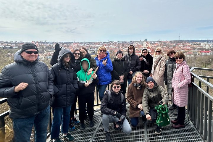Grupė suaugusiųjų ir vaikų stovi ant Altanos bokšelio terasos, nuo kurios atsiveria plati Vilniaus senamiesčio panorama. Dalyviai šiltai apsirengę, pozuoja šypsodamiesi – daugelis laiko nykščius į viršų ar mojuoja. Fone matyti žymūs Vilniaus architektūros