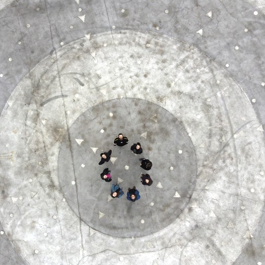 Nuotraukoje matomas betoninis paviršius su apskritimais ir geometriniais raštais, nufotografuotas iš viršaus. Centre stovi aštuoni žmonės, sustoję ratu ir žiūrintys į viršų.