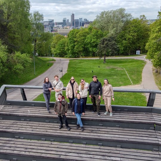 Grupė devynių žmonių stovi ant medinės lauko platformos, apsuptos žalios gamtos Vilniuje. Fone matyti miesto panorama su dangoraižiais ir debesuotu dangumi.
