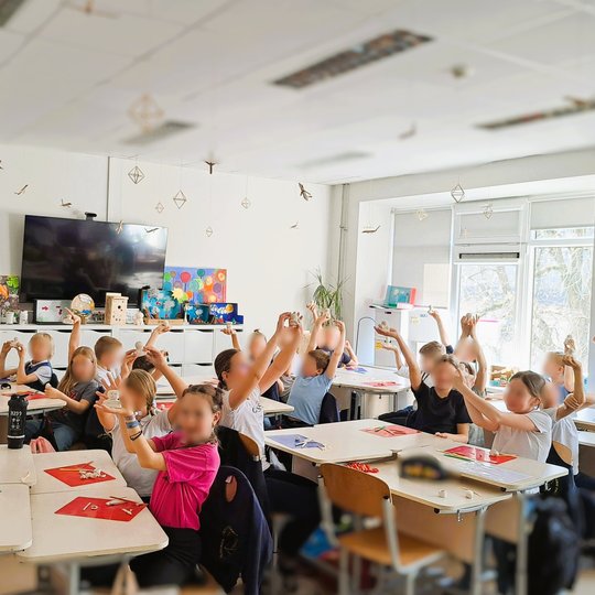 Nuotraukoje matome pradinių klasių mokinius klasėje. Vaikai sėdi prie stalų, pakėlę rankas aukštyn, laikydami kažką rankose – tai modelinas. Klasėje šviesu, pro didelius langus sklinda dienos šviesa. Ant sienų kabinami plakatai, vienas jų – „MŪSŲ GIMTADIE