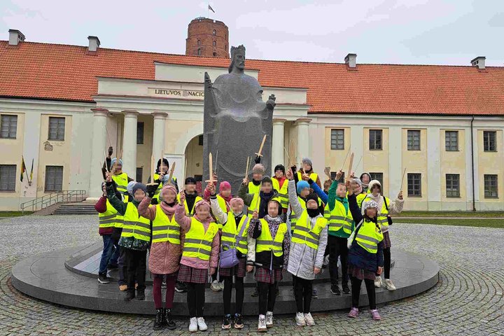 Grupė vaikų su ryškiais geltonais atšvaitiniais liemenėmis stovi prie Karaliaus Mindaugo paminklo Vilniuje, priešais Lietuvos nacionalinį muziejų, iškėlę medinius kalavijus; fone matyti Gedimino pilies bokštas.