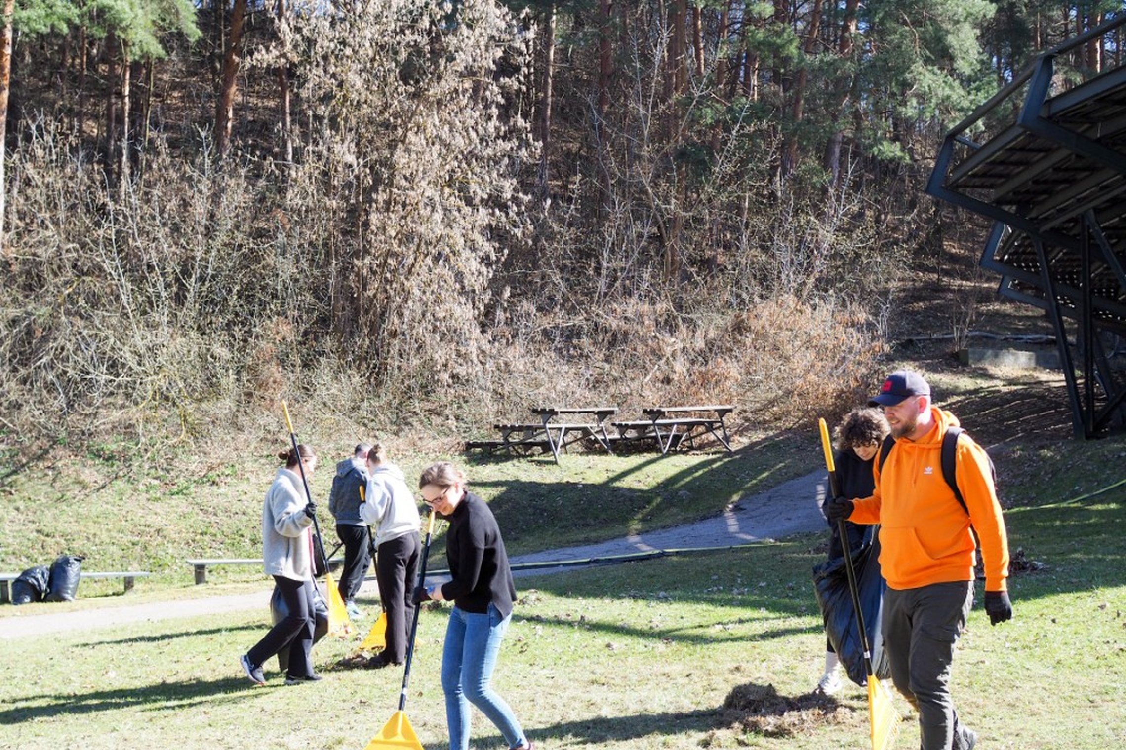 Grupė žmonių su grėbliais tvarko teritoriją lauke šalia miškingo šlaito. Saulėtas pavasario rytas, foną užpildo medžiai ir metalinė amfiteatro konstrukcija.