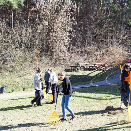 Grupė žmonių su grėbliais tvarko teritoriją lauke šalia miškingo šlaito. Saulėtas pavasario rytas, foną užpildo medžiai ir metalinė amfiteatro konstrukcija.