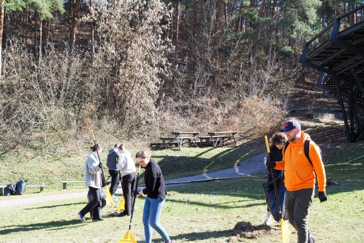 Grupė žmonių su grėbliais tvarko teritoriją lauke šalia miškingo šlaito. Saulėtas pavasario rytas, foną užpildo medžiai ir metalinė amfiteatro konstrukcija.