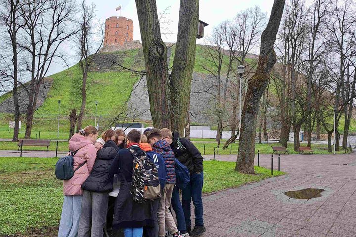 Grupė ketvirtokų stovi ratu ir susikaupę atlieka užduotį lauke prie Gedimino pilies kalno Vilniuje. Fone matoma Gedimino pilies bokštas ir žalias kalnas, apsuptas plikų medžių ir parko takų.