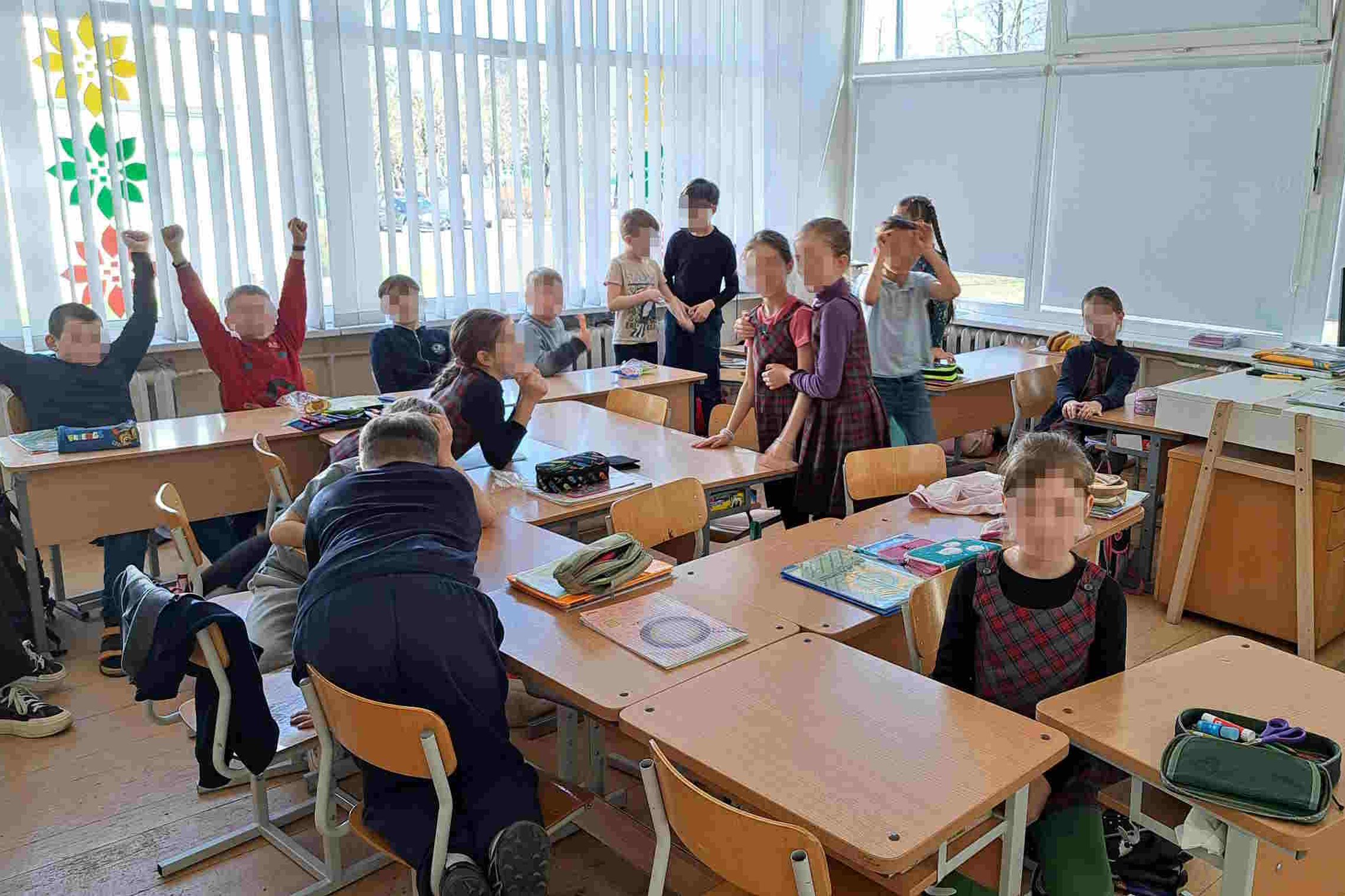 Klasės viduje susirinkusi vaikų grupė žaidžia ar dalyvauja aktyvioje veikloje. Kai kurie mokiniai stovi, kiti sėdi ar atsilošę, keli pakėlę rankas aukštyn. Ant stalų – sąsiuviniai, penalai, mokykliniai reikmenys. Klasės langai uždengti žaliuzėmis, ant jų 