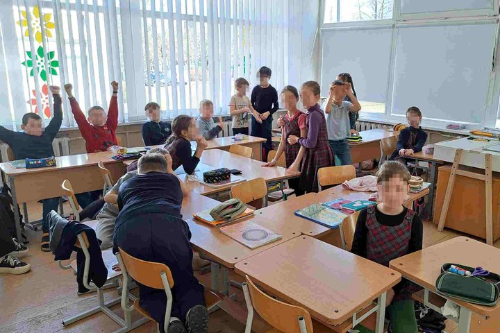 Klasės viduje susirinkusi vaikų grupė žaidžia ar dalyvauja aktyvioje veikloje. Kai kurie mokiniai stovi, kiti sėdi ar atsilošę, keli pakėlę rankas aukštyn. Ant stalų – sąsiuviniai, penalai, mokykliniai reikmenys. Klasės langai uždengti žaliuzėmis, ant jų 