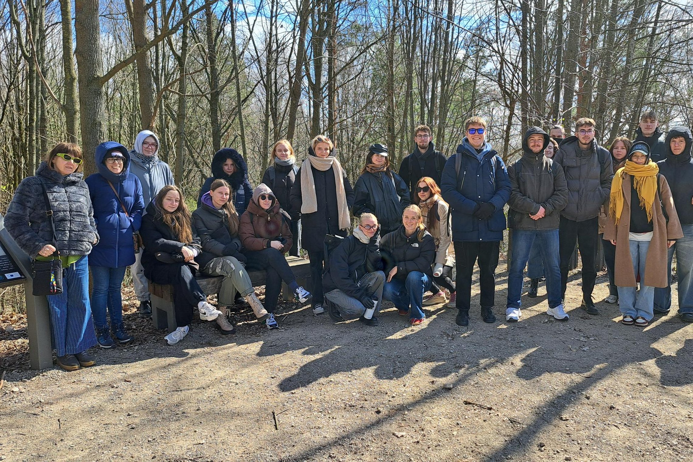 Ekskursijos grupė Vilniaus pilių teritorijoje. Žmonės įvairaus amžiaus ir lyties, apsirengę šiltai, stovi šaltą dieną. Atrodo žvarbu, nors šviečia saulė. Fone matyti plikos medžių lajos ir šviesiai mėlynas dangus.