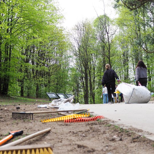 Prie miško tako ant žemės guli įvairūs darbo įrankiai, o fone žmonės neša didelį baltą maišą su surinktomis atliekomis. Šalia stovi dviratis ir matyti informaciniai stendai tarp žalios pavasarinės augmenijos.