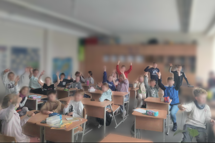 Trečios klasės mokiniai sėdi klasėje prie stalų, kai kurie atsistoję ar pakėlę rankas į viršų, šypsosi ir aktyviai dalyvauja veikloje. Klasės aplinka šviesi, lentynos pilnos priemonių, ant sienų kabo žemėlapiai ir plakatai. Vaikų veidai išplaukę dėl priva