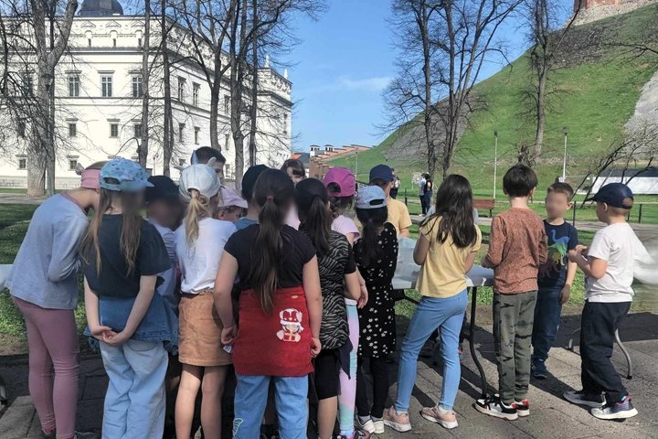 Grupė vaikų stovi susibūrę aplink stalą lauke, prie Gedimino pilies kalno Vilniuje. Fone matyti žalias kalno šlaitas su Gedimino bokštu ir Valdovų rūmai. Vaikai dėvi spalvingus drabužius, kepures, jų veidai atsukti į stalą, kur vyksta veikla. Dieną švieči