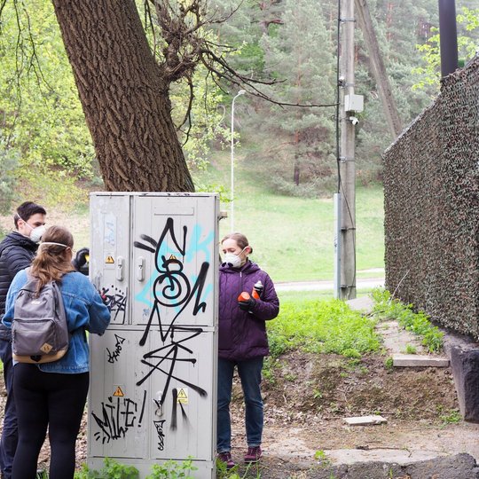 Keturi žmonės stovi prie elektros skydinės, ištepliotos grafičiais, ir ruošiasi ją valyti. Aplink matyti medžiai, žalia pieva ir pastatas, dengtas maskuojančia danga.