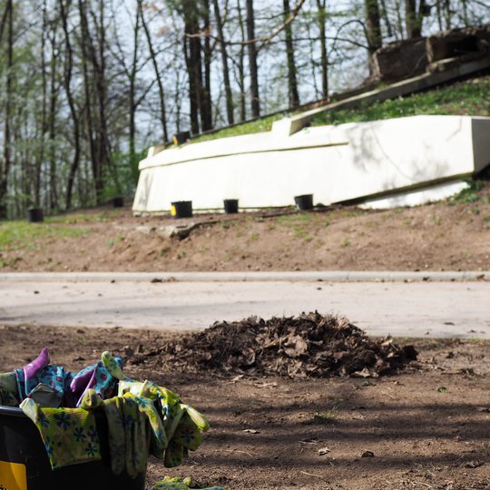 Kibiras su spalvingomis darbo pirštinėmis stovi šalia krūvos sugrėbtų lapų, tolumoje – baltas paminklas tarp medžių ir juodų vazonų.