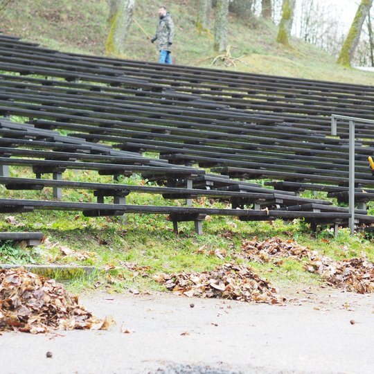 Vyras su mėlynu džemperiu ir kepure šluoja lapų krūvas lauko estradoje, šalia medinių žiūrovų suolų. Priekyje – kelių krūvų lapų eilė, o tolumoje matyti dar keli žmonės, atliekantys panašius darbus. Aplinka – miškinga, su pavasario žaluma.