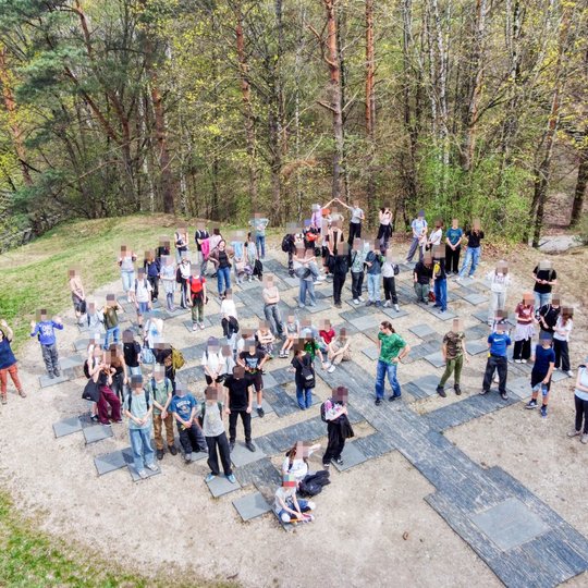 Šioje nuotraukoje matoma didelė grupė žmonių, stovinčių ant geneologinio medžio plytelių, lauko aplinkoje, apsuptoje medžių. Žmonės žvelgia į viršų ir mojuoja, nuotrauka daryta iš drono. Aplinka – pavasariška.