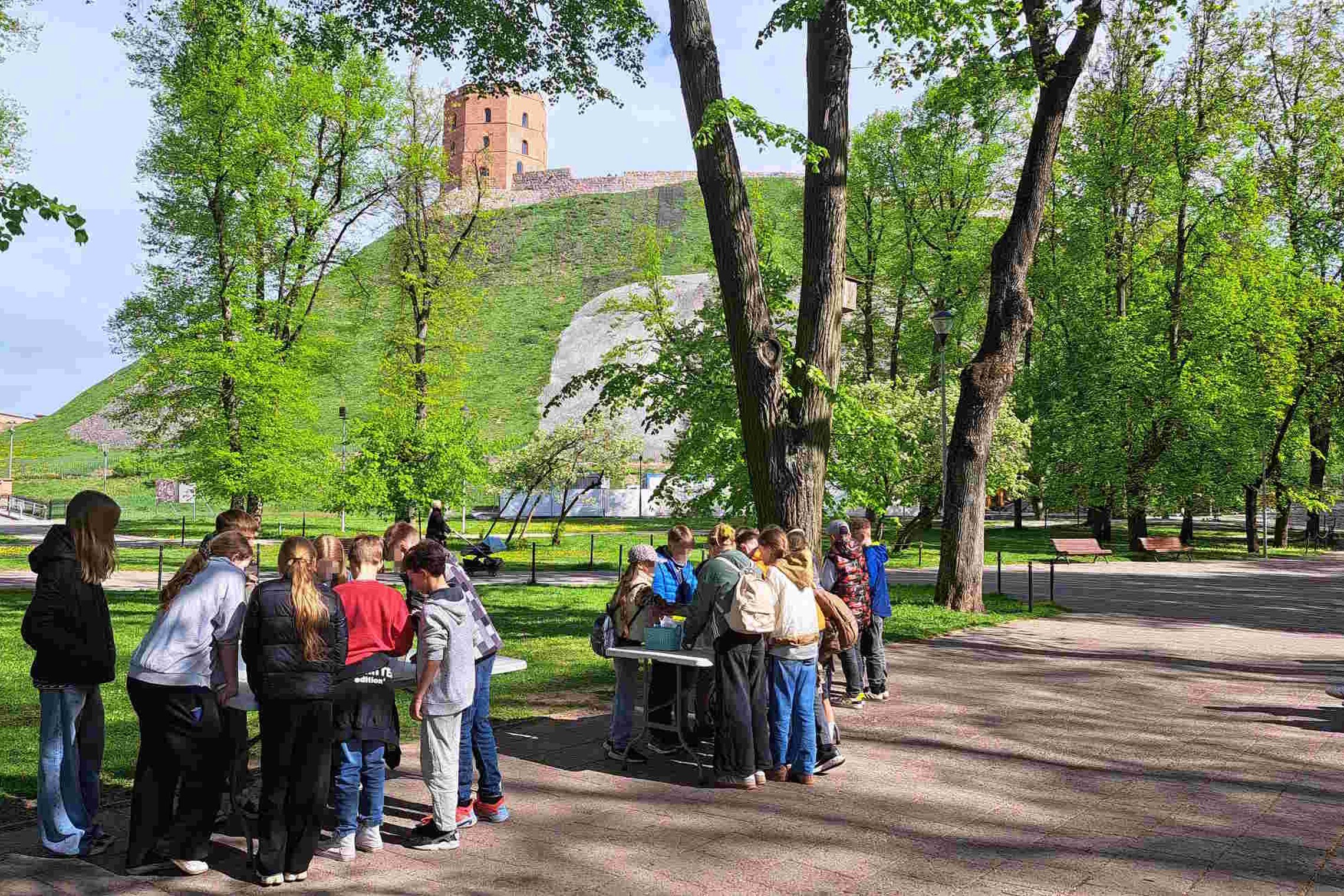 Pradinukai dalyvauja edukacinėje veikloje gryname ore šalia Gedimino pilies kalno Vilniuje. Vaikai, susiskirstę į grupes, tyrinėja informaciją prie stalų, apsupti žalumos ir pavasariškai sužaliavusių medžių.