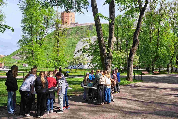 Pradinukai dalyvauja edukacinėje veikloje gryname ore šalia Gedimino pilies kalno Vilniuje. Vaikai, susiskirstę į grupes, tyrinėja informaciją prie stalų, apsupti žalumos ir pavasariškai sužaliavusių medžių.