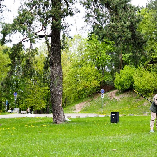 Moteris grėbia lapus žalioje pievoje, apsuptoje medžių ir kalvoto parko reljefo. Ji dėvi šiltus lauko drabužius, o šalia matyti eismo ženklai ir pėsčiųjų takas.