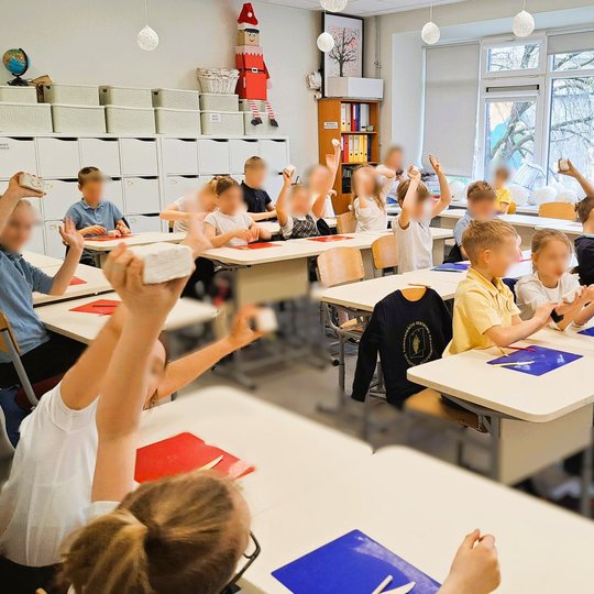Nuotraukoje matome pradinių klasių mokinius klasėje. Vaikai sėdi prie stalų, pakėlę rankas aukštyn, laikydami kažką rankose – tai modelinas. Klasėje šviesu, pro didelius langus sklinda dienos šviesa. Ant sienų kabinami plakatai, vienas jų – „MŪSŲ GIMTADIE