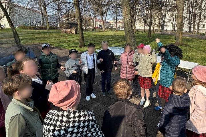 Antros klasės mokiniai lauke dalyvauja edukacijoje Vilniaus pilių rezervato teritorijoje, fone matyti Valdovų rūmai. Vaikai susibūrę ratu nuotaikingai diskutuoja, o rato viduryje matyti mergaitė iškėlusi rankas į orą, kuri dėvi panko peruką.