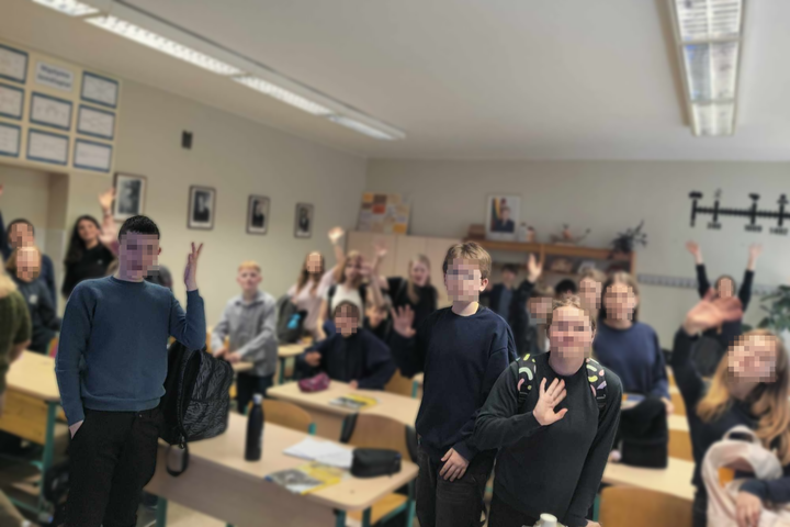 Mokiniai klasėje linksmai mojuoja po edukacinės pamokos apie Vilniaus pilių istoriją. Priekyje stovi keli mokiniai, o kiti sėdi ar stovi prie savo suolų. Klasėje jauki aplinka, ant sienų – portretai ir lentelės su mokyklos informacija.