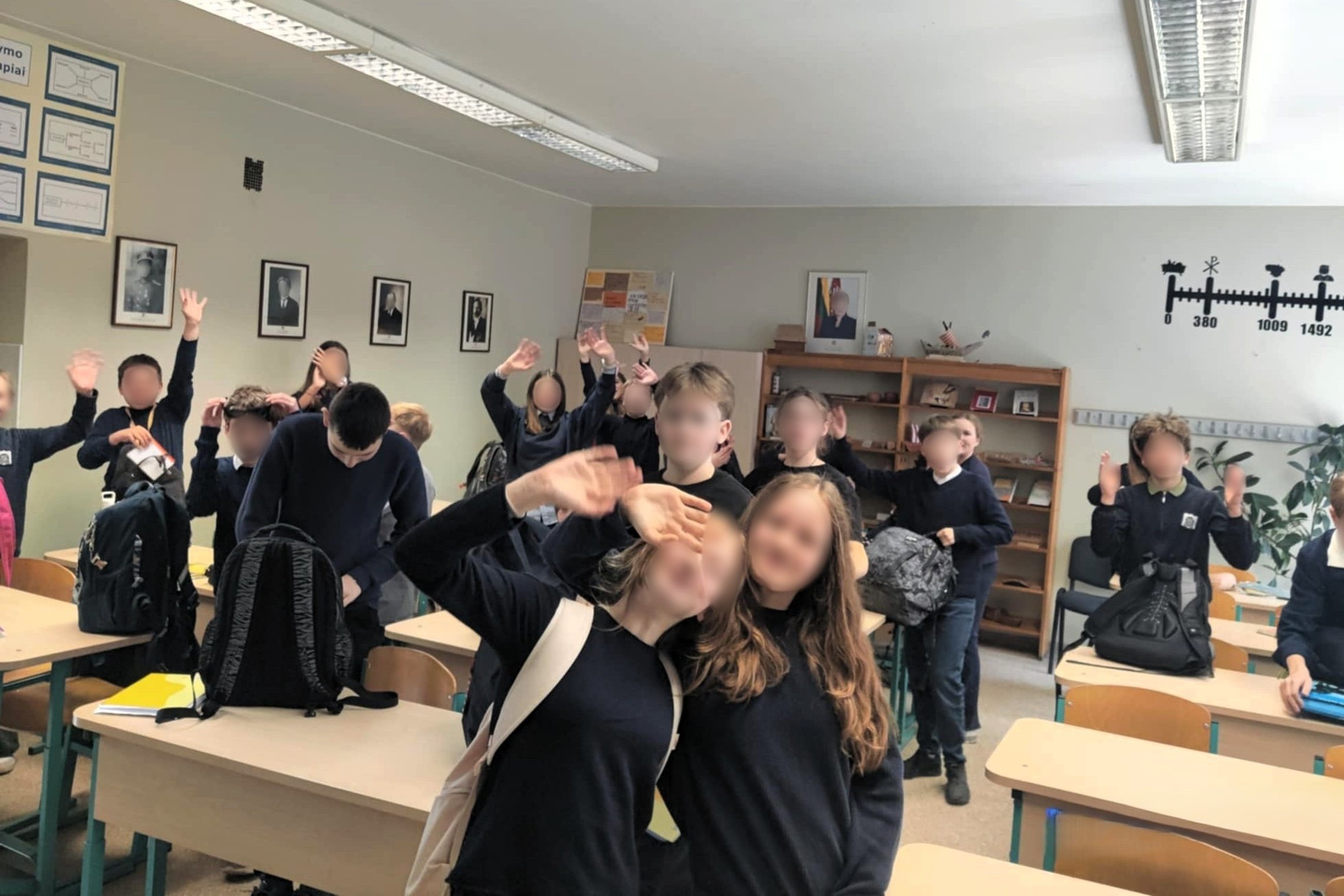 Klasėje susirinkę mokiniai mojuoja ir šypsosi. Matyti šviesi klasės aplinka su istorijos laiko juosta ant sienos ir nuotraukomis bei lentynomis fone.