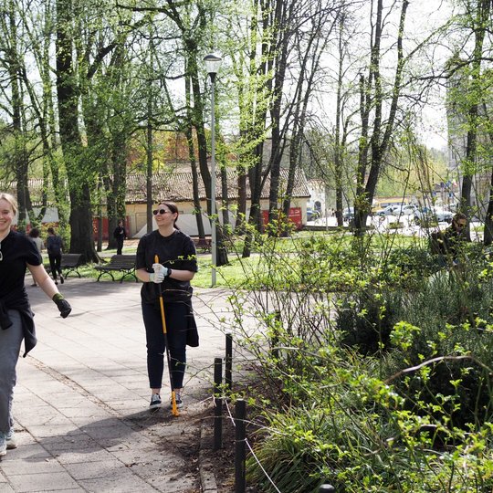 Šioje nuotraukoje matome grupę žmonių, dalyvaujančių aplinkos tvarkymo talkoje. Priekyje – dvi moterys, viena iš jų neša šiukšlių maišą, kita – laiko grėblį. Jos šypsosi, o aplinka žaliuoja – matosi daug medžių su šviežiai išsiskleidusiais lapais. Tai gre